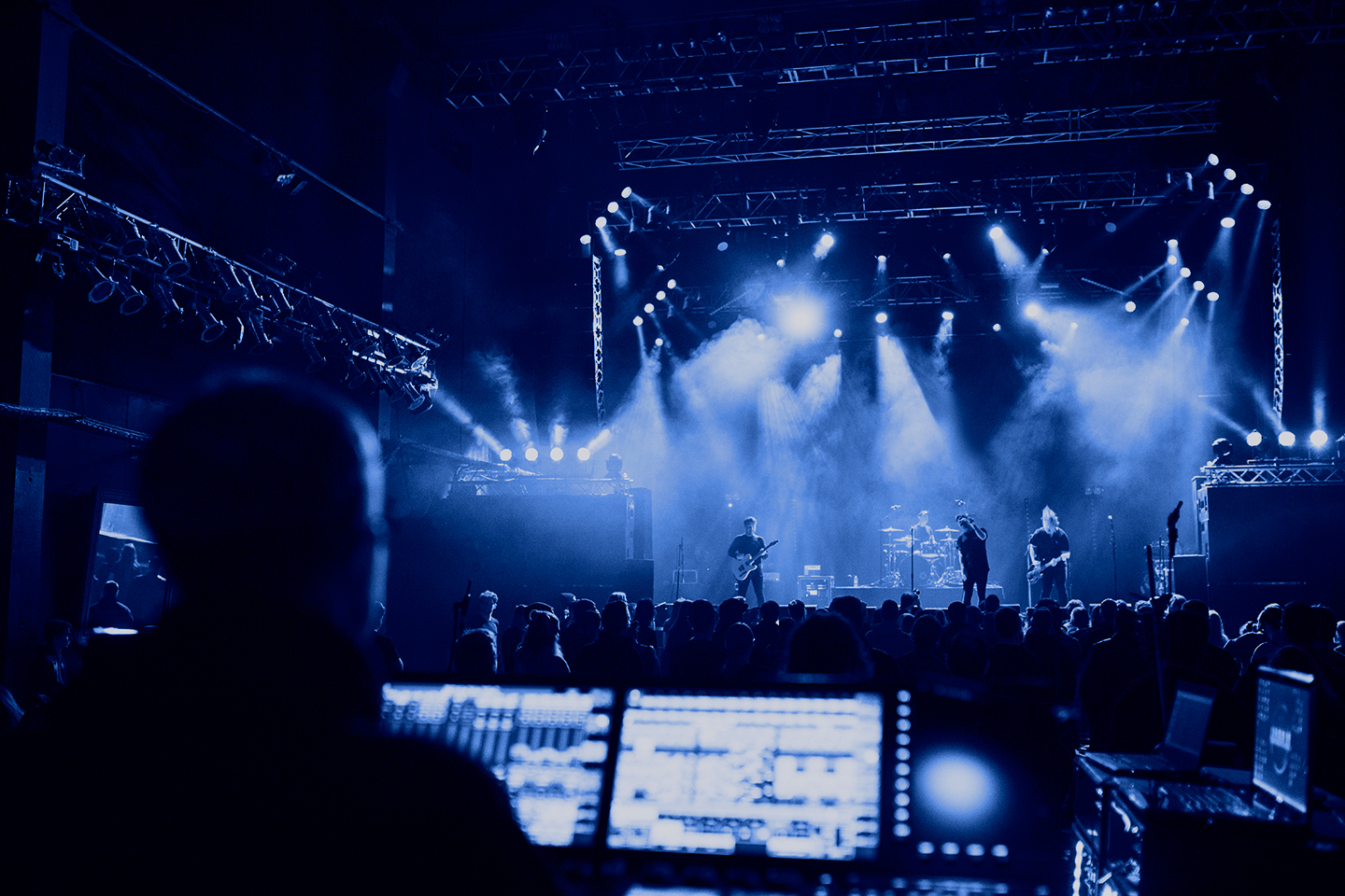 Front-of-house perspective at a live concert, mixing consoles in the foreground and stage lights over the audience